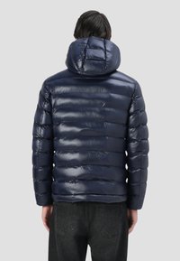 Giubbotto imbottito blu navy con finitura lucida, caratterizzato da sezioni trapuntate orizzontali e un cappuccio, indossato sopra jeans scuri.