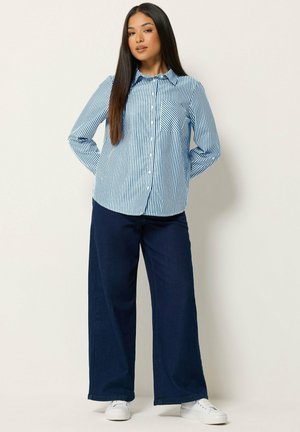 Frau mit langen dunklen Haaren, die ein blau-weiß gestreiftes Hemd, dunkle weite Hosen und weiße Turnschuhe trägt, steht mit den Händen hinter dem Rücken.