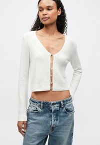 Mujer vistiendo un cárdigan blanco corto con botones dorados y jeans azules holgados, de pie contra un fondo claro y liso.
