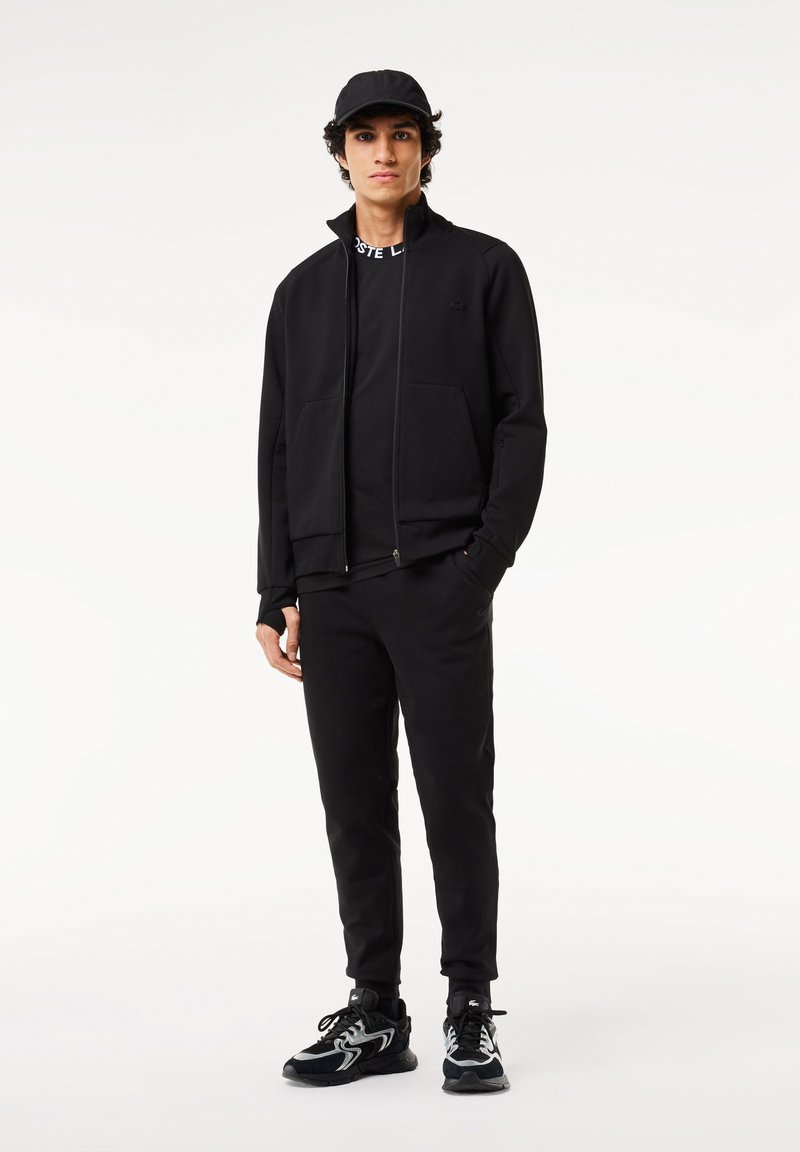 Completo sportivo nero con giacca con zip e tasche, abbinato a pantaloni affusolati coordinati. Sneakers nere con accenti grigi.