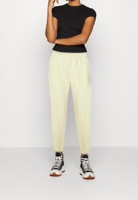 Světlé žluté tepláky s elastickým pasem a manžetami, se bočními pruhy a logem. Doplňuje je černé vysoké tenisky.