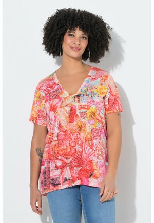 Frau mit lockigem Haar, die große Creolenohrringe trägt und ein buntes V-Ausschnitt-Shirt mit pinken und orangen Mustern vor einer weißen Wand steht.