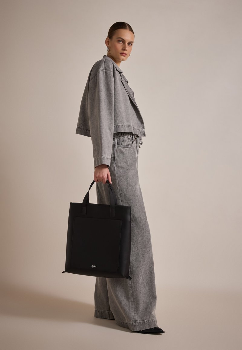 Sac fourre-tout en cuir noir avec deux poignées courtes, présentant une poche avant. Le modèle porte une veste en denim grise à manches courtes et un pantalon large.