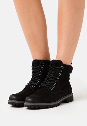 Botas de tobillo de ante negro con un collar acolchado, puntera redonda y cordones negros. Suela robusta con surcos profundos para mayor tracción.