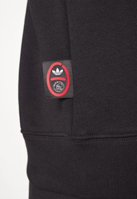 Svart sweatshirt-tyg med ribbad textur. Har en svart etikett med röda detaljer som visar en logotyp med tre ränder och en grafisk design.