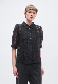 Blusa nera in pizzo con maniche corte e collo a camicia, caratterizzata da intricati motivi floreali e chiusura frontale con bottoni, abbinata a pantaloni scuri.