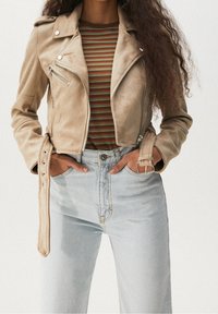 Veste biker courte en daim beige avec poches zippées, associée à une chemise rayée multicolore et un jean taille haute en denim clair.