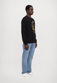 Felpa nera con grafica di un serpente dorato, colletto rotondo, maniche lunghe e polsini a coste, abbinata a jeans azzurri chiari e sneakers bianche.