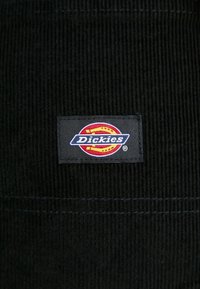 Μαύρος βελουτέ ύφασμα με ορθογώνια μαύρη πινακίδα που διαθέτει το πολύχρωμο λογότυπο "Dickies". Ορατές απότομες ραβδώσεις σε όλο το υλικό.