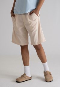 Beige tailored shorts with side pockets and a relaxed fit, paired with white ribbed socks and brown clogs, set against a plain background.