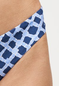 Κοντινή λήψη υφάσματος με γεωμετρικό σχέδιο σε μπλε και ανοιχτό μπλε στην περιοχή του ισχίου ενός ατόμου με μαυρισμένο δέρμα.