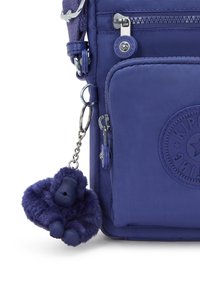 Lila nylon crossbody-väska med en texturerad yta, blixtlåsfack och en fluffig lila apa nyckelring fäst.
