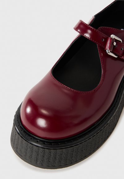 Burgundy lackskinn Mary Jane-sko med rund tå, justerbar spänne och tjock svart plattformssula med en texturerad yta.