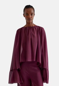 Blusa fluida bordeaux con design drappeggiato, scollo arricciato e maniche lunghe a campana. Realizzata in tessuto leggero con una texture morbida.