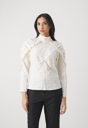 Blusa bianca a maniche lunghe con accentuazioni arricciate sulle spalle, superficie testurizzata, collo alto e dettagli arricciati al centro davanti.