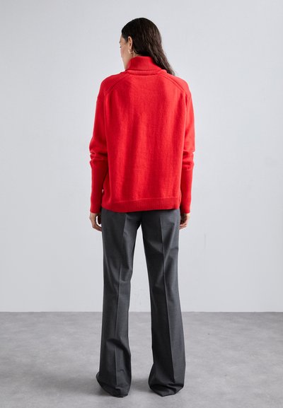 Red knitted sweater with a high collar, ribbed cuffs, and hem, worn over grey tailored trousers with a straight leg and slight flare.