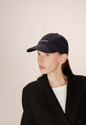 Casquette de baseball en coton bleu marine avec logo brodé, visière incurvée et strap ajustable. Portée avec un blazer noir et un top blanc.