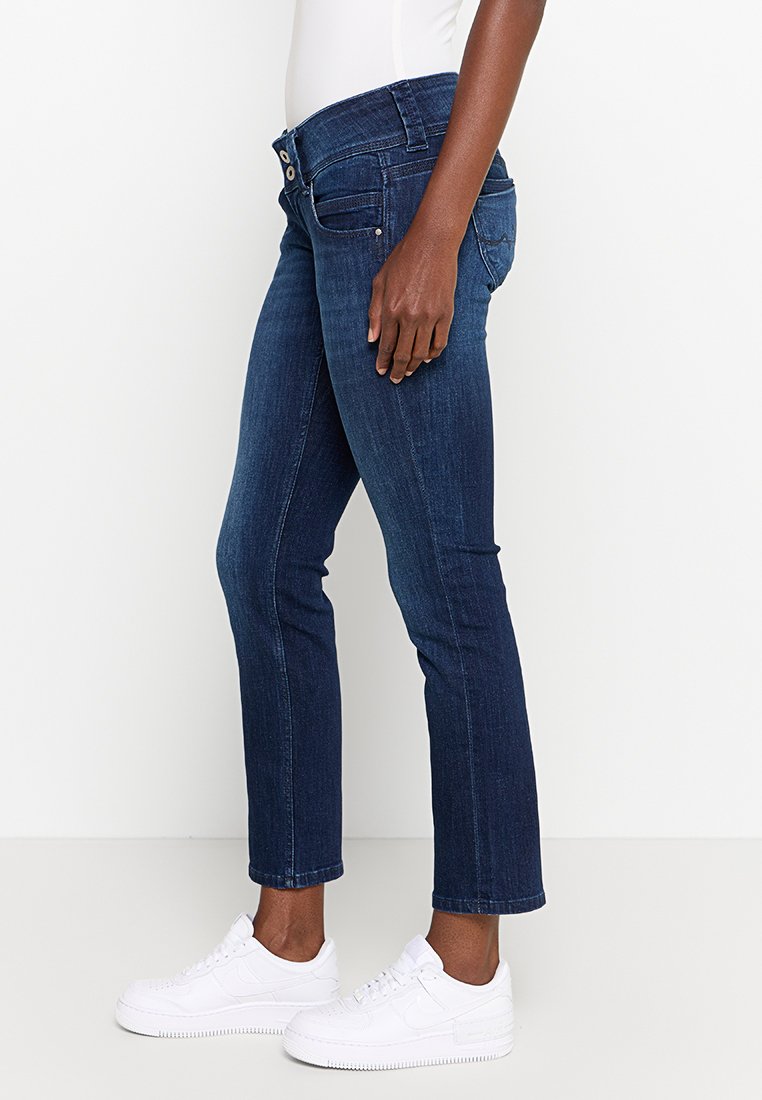 Jeans de mezclilla azul oscuro con un diseño ajustado, cintura de altura media, dos bolsillos frontales y corte recto, combinados con zapatillas deportivas blancas.