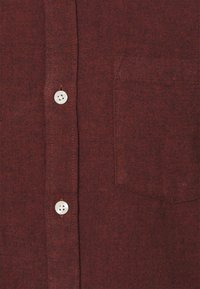 Chemise boutonnée bordeaux en tissu texturé, avec une poche poitrine et des boutons blancs, affichant un motif tissé.