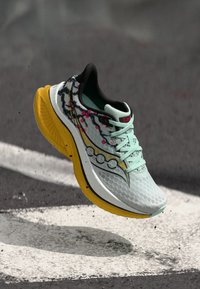 Ανοιχτό μπλε παπούτσι για τρέξιμο με κίτρινη σόλα, μαύρο και ροζ φλοράλ σχέδιο, στον αέρα πάνω από άσφαλτο με λευκή οδική σήμανση.