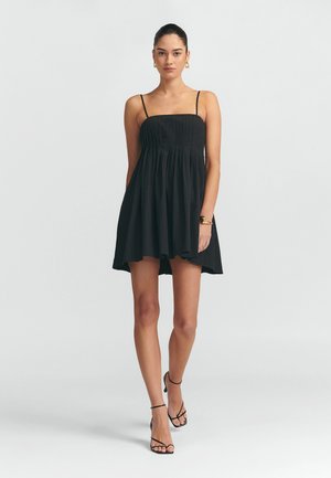 Robe noire avec un corsage ajusté, texture plissée et jupe évasée. Fines bretelles et longueur au-dessus du genou, associée à des talons noirs à lanières.