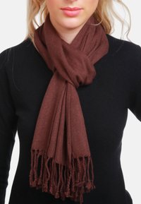 Écharpe marron tissée avec des bords effilochés, enroulée autour du cou, en matériau doux, tombant doucement sur un haut noir.