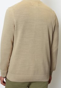 Homme portant un pull en maille beige à manches longues et un pantalon vert olive, vu de dos sur un fond blanc uni.