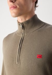 Maglione zip-up verde oliva con colletto alto, texture a coste e una toppa rossa con il marchio "HUGO" sul lato sinistro.