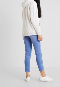 Osoba v světle šedé mikině s kapucí, modrých úzkých džínách a bílých teniskách stojící před jednoduchým světle šedým pozadím.