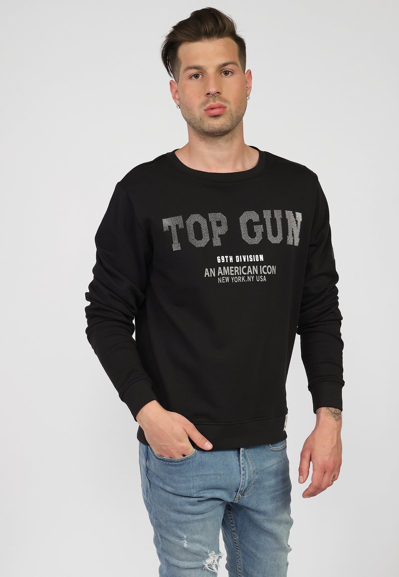 Czarny sweatshirt z napisem "TOP GUN" w fakturowanych szarych literach, okrągły dekolt, długie rękawy i ściągacze na mankietach. Noszony z podartymi jasnoniebieskimi dżinsami.