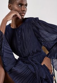 Granatowa plisowana sukienka z długimi, rozkloszowanymi rękawami i paskiem w talii. Teksturowana tkanina z subtelnym połyskiem. Szczegół w falbance na dole.