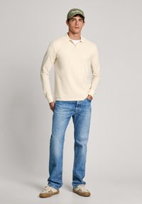 Ilgi piedurkņu bēšs polo krekls, zilas džinsu bikses un bēšas sporta apavi. Pelēka cepure ar baltu logotipu. Modelis stāv, rokas kabatās, neitrāls fons.