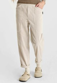Beige corduroy pants with a relaxed fit, featuring two front pockets, visible seam details, and rolled cuffs. Styled with clogs and patterned socks.