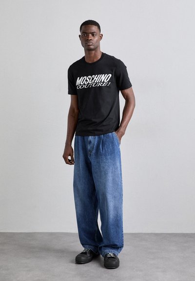 T-shirt nera in cotone con stampa bianca "MOSCHINO COUTURE!", abbinata a jeans a forma libera in denim blu e sneakers nere.