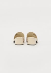 Mules à talons beiges avec un dessus en cuir lisse, une arrière ouvert et un bout carré. Dotés d'un talon épais de couleur noire contrastante.