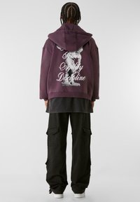 Sudadera morada con un estampado gráfico en la parte posterior que dice "Poder Agilidad Disciplina", combinada con pantalones cargo negros y zapatillas blancas.