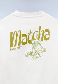 Λευκό πουκάμισο με το κείμενο «Matcha Since 1987» και καρτούν χαρακτήρα ποτού matcha που κρατάει μια πινακίδα με τη λέξη «Πρεσβευτής» σε πράσινο χρώμα.