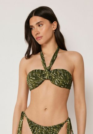 Femme aux longs cheveux foncés portant un haut de bikini licou tie-dye vert olive et un bas de bikini assorti avec des liens sur les côtés, sur un fond uni.
