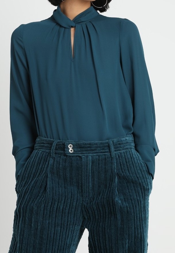 Femme portant un chemisier manches longues bleu sarcelle foncé avec un décolleté goutte d'eau et un pantalon en velours côtelé bleu sarcelle foncé taille haute, mains dans les poches.
