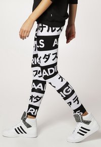 Osoba jde oblečena v černo-bílých legínách se výraznými vzory japonského a anglického textu, doplněná bílými vysokými teniskami Adidas.