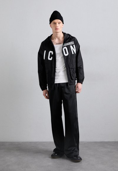Schwarze Windjacke mit großen weißen Buchstaben "ICON" und Markenlogo, kombiniert mit einem weißen Tanktop und lockeren schwarzen Hosen. Strickmütze in Schwarz.