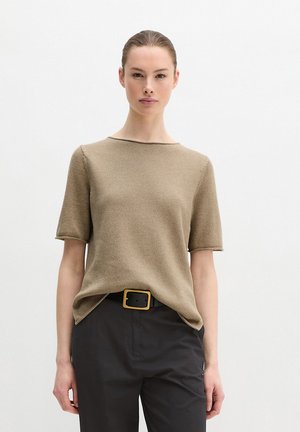 Frau trägt ein beige kurzärmeliges Strickoberteil, in schwarze Hosen gesteckt, mit einem schwarzen Gürtel, der eine große quadratische goldene Schnalle hat.