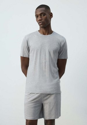 Hombre de pie con las manos detrás de la espalda, vistiendo una camiseta de manga corta lisa de color gris claro y pantalones cortos a juego sobre un fondo neutro.