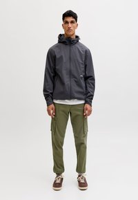 Giovane in piedi rivolto in avanti che indossa una giacca con cappuccio grigio scuro, pantaloni cargo verde oliva e scarpe da ginnastica marroni su sfondo bianco uniforme.