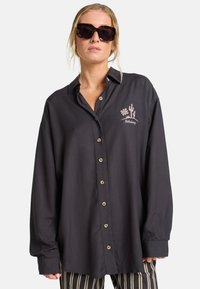 Camisa oscura de corte holgado con botones en el frente, que presenta un bordado de cactus y flores en color rosa claro. El material parece suave.