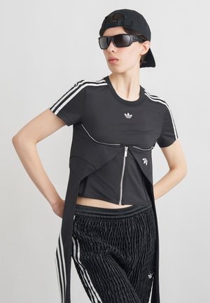 Osoba pózuje v černém sportovním oblečení Adidas s bílými pruhy, nosí velké sluneční brýle a černou baseballovou čepici otočenou dozadu na jednobarevném pozadí.