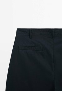 Arrière d'un pantalon noir avec passants de ceinture et une poche passepoilée horizontale sur le côté droit sur fond blanc.