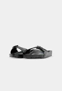 Paire de sandales noires à enfiler avec une seule bride ajustable et des semelles texturées sur un fond blanc uni.