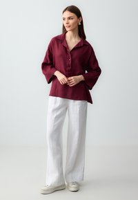 Bordowa lniana koszula z kołnierzykiem i guzikami, w zestawieniu z luźnymi białymi lnianymi spodniami. Ubranie ma swobodny krój i teksturowaną tkaninę.