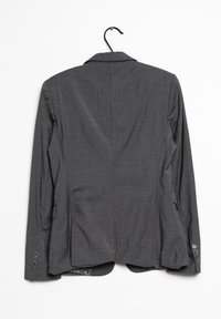 Grijze blazer van gestructureerde stof, met een reverskraag, een achterkant met een split en zes knopen op de manchetten. Hangt aan een zwarte haak.